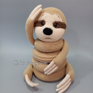 Könnte beinhalten: Ein gehäkeltes Faultier-Spielzeug mit braunem Fell, einer schwarzen Nase und braunen Augen. Das Faultier sitzt mit seinen Armen um seinen Körper gewickelt und seinen Kopf zur Seite geneigt. Das Spielzeug ist aus Garn gefertigt und etwa 25 cm groß.