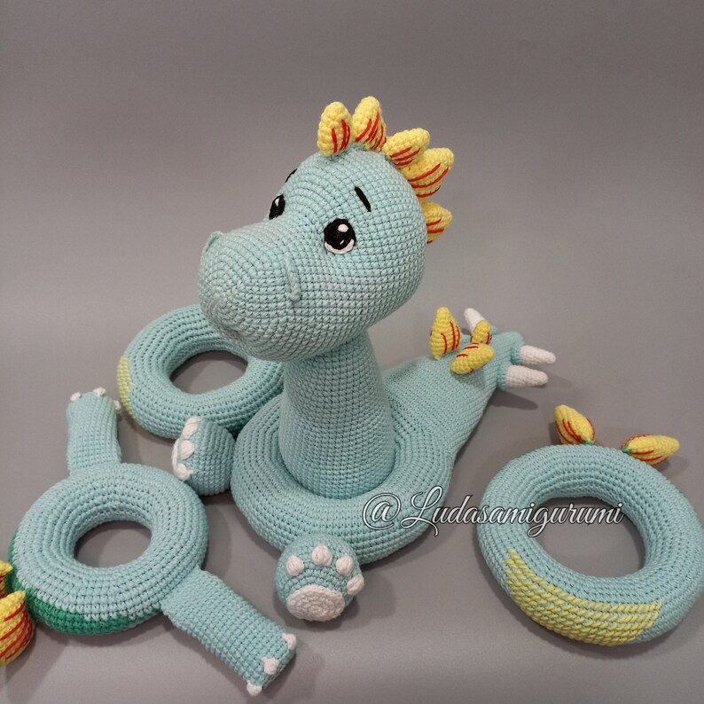 May include: A crocheted turquoise dinosaur toy with yellow and red accents. The dinosaur has three rings attached to it, also crocheted in turquoise with yellow accents. The rings are for a baby to play with.