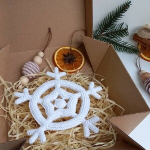 May include: A white crocheted snowflake ornament in a brown cardboard box with shredded paper packing. The box is open and the snowflake is visible.