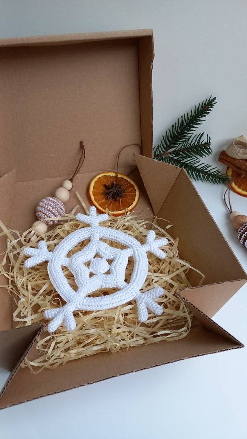 May include: A white crocheted snowflake ornament in a brown cardboard box with shredded paper packing. The box is open and the snowflake is visible.
