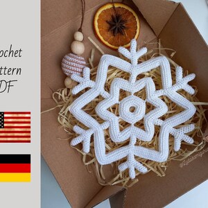 May include: A white crochet snowflake, displayed in a cardboard box with dried orange slices and decorative wooden beads. The text "Crochet pattern PDF" is visible, along with the American and German flags.