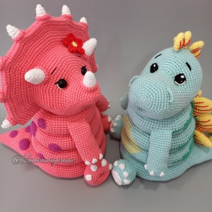May include: Two crocheted stuffed dinosaur toys. The triceratops is pink with purple spots and a red flower on its head. The other dinosaur is light blue with yellow accents and a yellow and orange crest.
