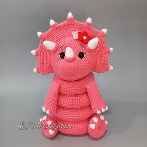 May include: A pink crocheted triceratops stuffed animal with white horns and a white flower on its head. The triceratops is sitting on a gray surface.