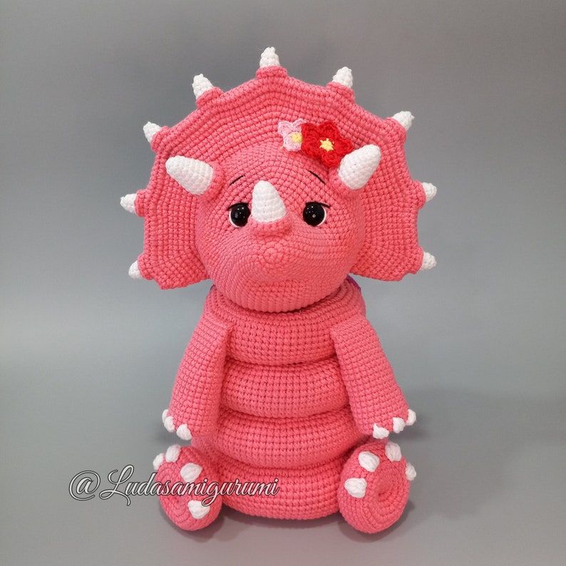 May include: A pink crocheted triceratops stuffed animal with white horns and a white flower on its head. The triceratops is sitting on a gray surface.