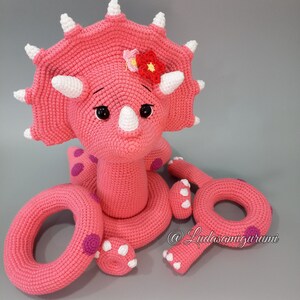 May include: Pink crocheted triceratops stuffed animal with a flower on its head. The triceratops is sitting on a pink crocheted ring and has two smaller pink crocheted rings beside it.