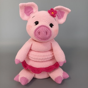 May include: A handmade crochet pig doll in shades of pink. The pig has a flower on its head and a ruffled skirt. The pig's ears, snout, and feet are a darker shade of pink. The doll is sitting with its legs extended.
