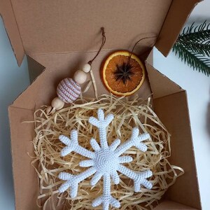 May include: A white crocheted snowflake ornament, a brown wooden bead garland with a white and brown striped bead, and a dried orange slice with a star anise inside, all packaged in a brown cardboard box with shredded paper.