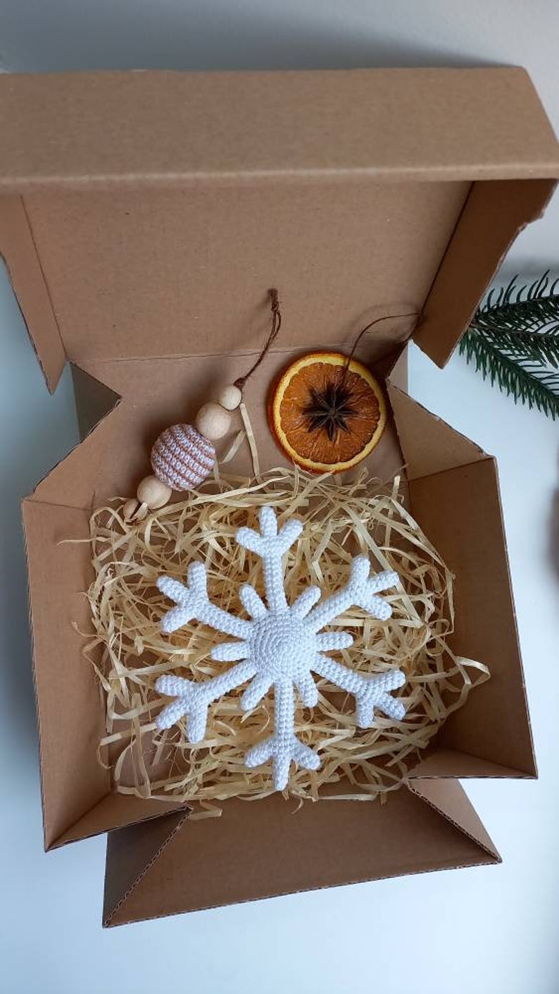 May include: A white crocheted snowflake ornament, a brown wooden bead garland with a white and brown striped bead, and a dried orange slice with a star anise inside, all packaged in a brown cardboard box with shredded paper.