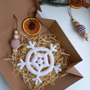 May include: A white crocheted snowflake ornament in a brown cardboard box with shredded paper. The box also contains a brown wooden bead garland with a brown and white striped bead, a dried orange slice, and a cinnamon stick with a brown ribbon.
