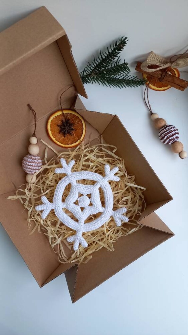 May include: A white crocheted snowflake ornament in a brown cardboard box with shredded paper. The box also contains a brown wooden bead garland with a brown and white striped bead, a dried orange slice, and a cinnamon stick with a brown ribbon.