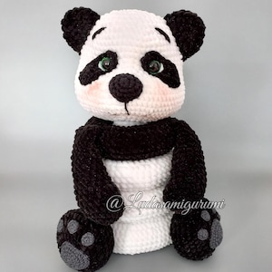 May include: A crocheted panda bear plush toy with black and white fur. The panda has a black nose, green eyes, and gray paw pads. The toy is sitting on a gray background. @Ludasamigurumi
