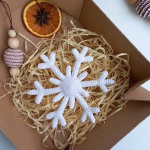 May include: A white crocheted snowflake ornament in a brown cardboard box with shredded paper. The box also contains a dried orange slice and a crocheted ball ornament with wooden beads.