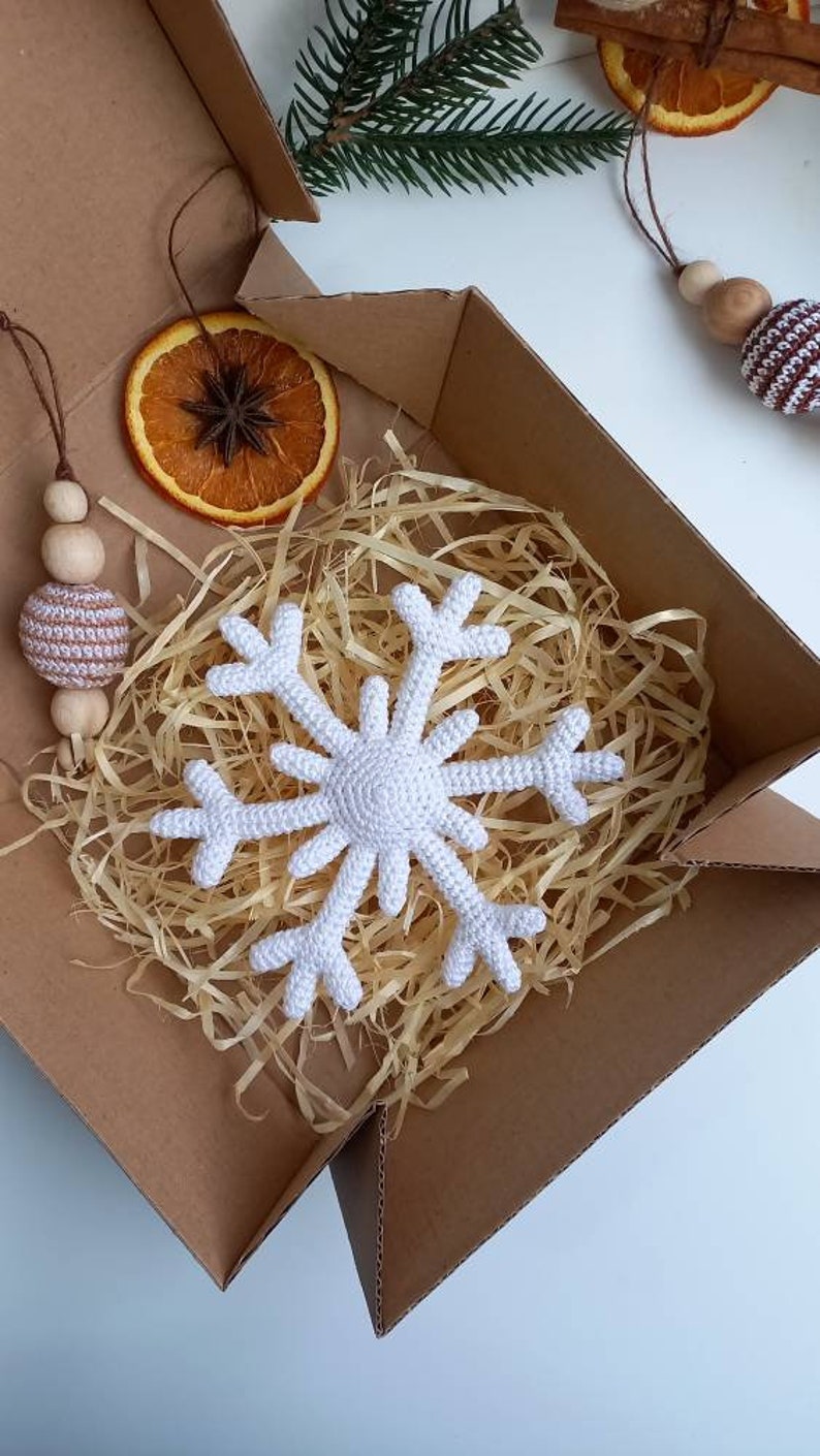 May include: A white crocheted snowflake ornament in a brown cardboard box with shredded paper. The box also contains a dried orange slice and a crocheted ball ornament with wooden beads.