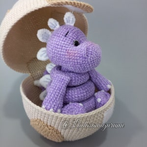 Może przedstawiać: Fioletowy, szydełkowy smok siedzący w szydełkowym jajku. Jajko jest otwarte, a smok patrzy w prawo. Smok ma białe, szydełkowe kolce na grzbiecie i głowie. Jajko jest beżowe i brązowe z białymi akcentami. Na jajku widoczny jest tekst "@Ludasamigurumi".