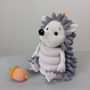 Puede incluir: Un juguete de erizo de punto de crochet gris y blanco con ojos negros y nariz marrón. El erizo está sentado en una superficie gris con una pequeña bellota de crochet a su lado. El juguete está hecho por @Ludasamigurumi.
