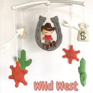 Puede incluir: Un móvil de bebé de crochet con un vaquero, cactus, estrellas y una bolsa de dinero. El móvil es blanco y el vaquero lleva un sombrero marrón y una bandana roja. El texto "Wild West Crib Mobile Pattern" está debajo del móvil.