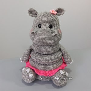 May include: A gray crocheted hippopotamus wearing a pink tutu and a pink flower on its head. The hippopotamus has black eyes and pink cheeks.