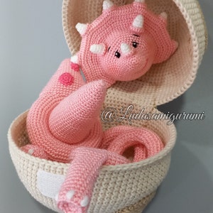 May include: A pink crocheted dinosaur toy in a crocheted egg-shaped basket. The dinosaur has a long tail and three horns on its head. The basket is beige and has a white label on the side. The text "@Ludasamigurumi" is visible on the basket.