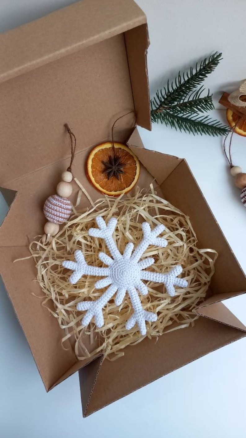 May include: A white crocheted snowflake ornament in a brown cardboard box with shredded paper, a dried orange slice, and a brown and white crocheted ball ornament.