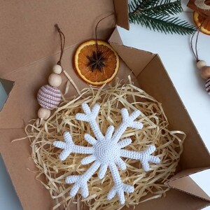 May include: A white crocheted snowflake ornament in a brown cardboard box with shredded paper, a dried orange slice, and a brown and white crocheted ball ornament.