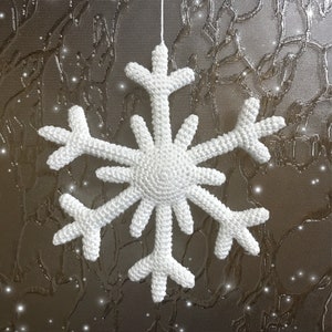 May include: A white, crocheted snowflake ornament suspended by a thin white string. The snowflake has six symmetrical arms, each with multiple points. The background is a textured, metallic surface with a subtle pattern and sparkling highlights.