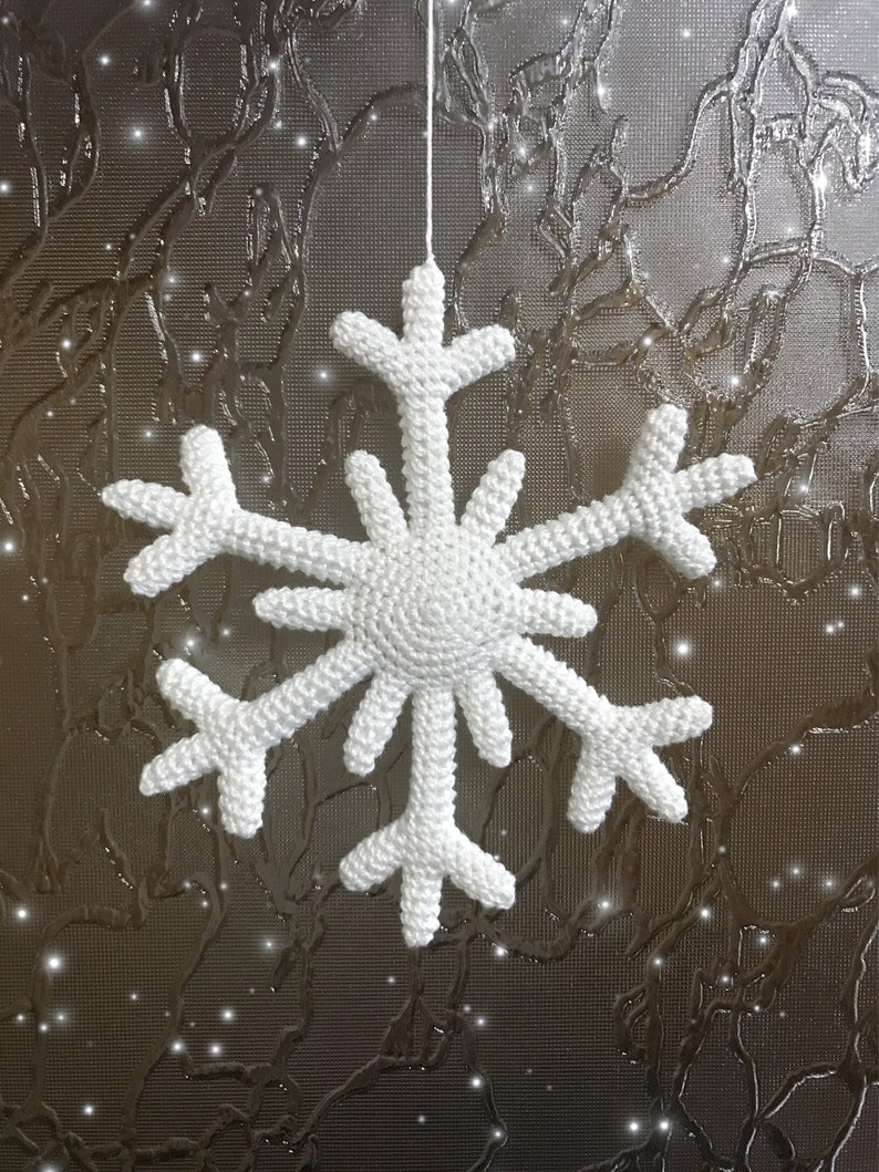 May include: A white, crocheted snowflake ornament suspended by a thin white string. The snowflake has six symmetrical arms, each with multiple points. The background is a textured, metallic surface with a subtle pattern and sparkling highlights.