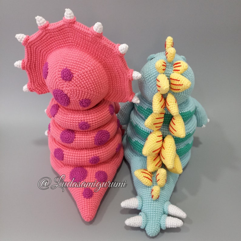 May include: Two crocheted dinosaur toys, one pink with purple spots and a frill, the other blue with yellow stripes and a spiky back.  @Ludasamigurumi