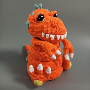 May include: An orange crochet dinosaur toy with white teeth, white claws, and blue accents. The handmade stuffed animal has large black eyes and is sitting upright. The dinosaur's claws have yellow tips.