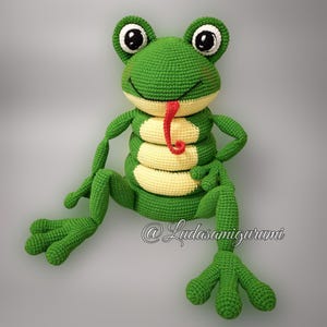 Puede incluir: Una rana de crochet verde con la lengua roja que sobresale. La rana tiene grandes ojos negros y una barriga amarilla. La rana está sentada sobre un fondo gris. @Ludasamigurumi