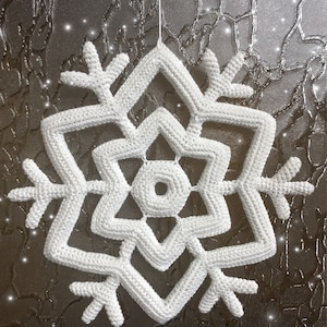 May include: A white, crocheted snowflake ornament. The snowflake is suspended by a thin white string. The design features a six-pointed star shape with intricate detailing. The background is a textured, silver-toned surface with sparkling accents.