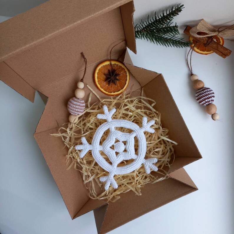 May include: A white crocheted snowflake ornament in a brown cardboard box with shredded paper. The box is open and there are two other ornaments, a brown and white crocheted ball and a wooden ball with a brown and white crocheted ball on top, visible in the background.