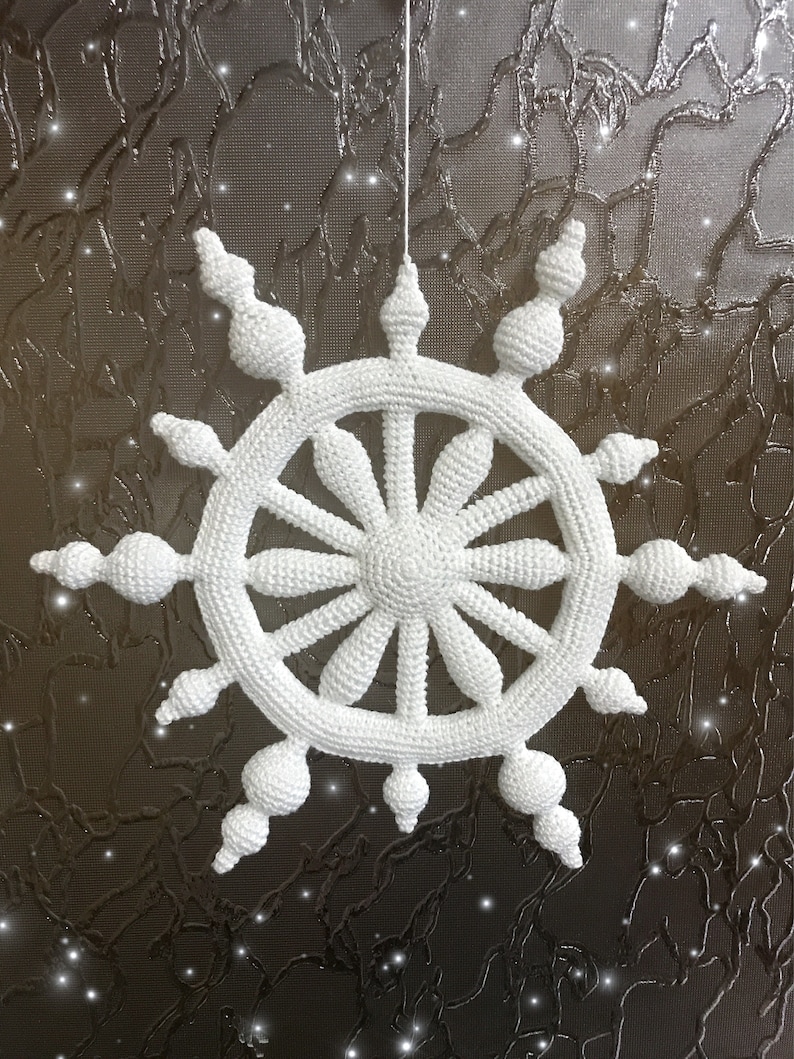 May include: A white, crocheted snowflake ornament. The snowflake is suspended by a white string and features a circular design with radiating spokes and decorative, rounded elements. The background is a textured, dark surface.