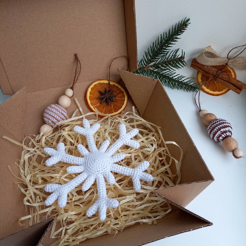 May include: A white crocheted snowflake ornament in a cardboard box with brown packing paper. The snowflake is surrounded by brown packing paper. There are other ornaments in the box, including a brown and white striped ornament and a dried orange slice.