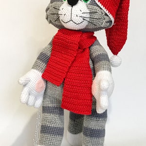 Może przedstawiać: Szary i biały szydełkowy kot w czerwonym szydełkowym kapeluszu i szaliku. Kot ma zielone oczy i biały nos. Kot stoi na białym tle.