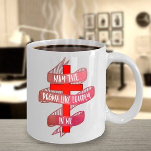 May include: White ceramic coffee mug with a red cross and pink and white ribbon with the text "May the dogma live loudly in me".