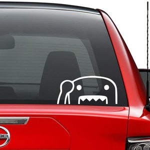 May include: White vinyl decal of a cartoon monster with a toothy grin, waving with one arm. The decal is applied to the rear window of a red car.