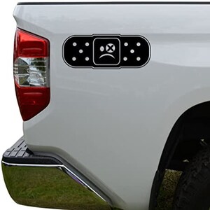 May include: A black and white vinyl decal of a band-aid with a sad face and an X over the eye. The decal is on the back of a white pickup truck.