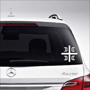 May include: White vinyl decal of a cross with the letters "DC" on each arm. The decal is on the back window of a silver car.