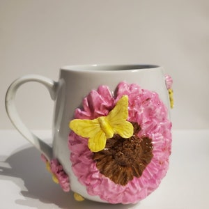 Puede incluir: Taza de cerámica blanca con una flor rosa y marrón y una mariposa amarilla esculpida en el lateral.