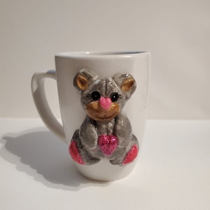 Puede incluir: Taza de cerámica blanca con un oso de peluche gris que sostiene un corazón rosa. El oso tiene orejas marrones y una nariz rosa.