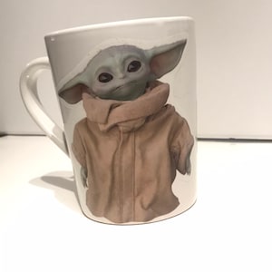 Puede incluir: Taza de cer&aacute;mica blanca con una imagen colorida de un personaje de la franquicia Star Wars, The Mandalorian, conocido como "Baby Yoda".