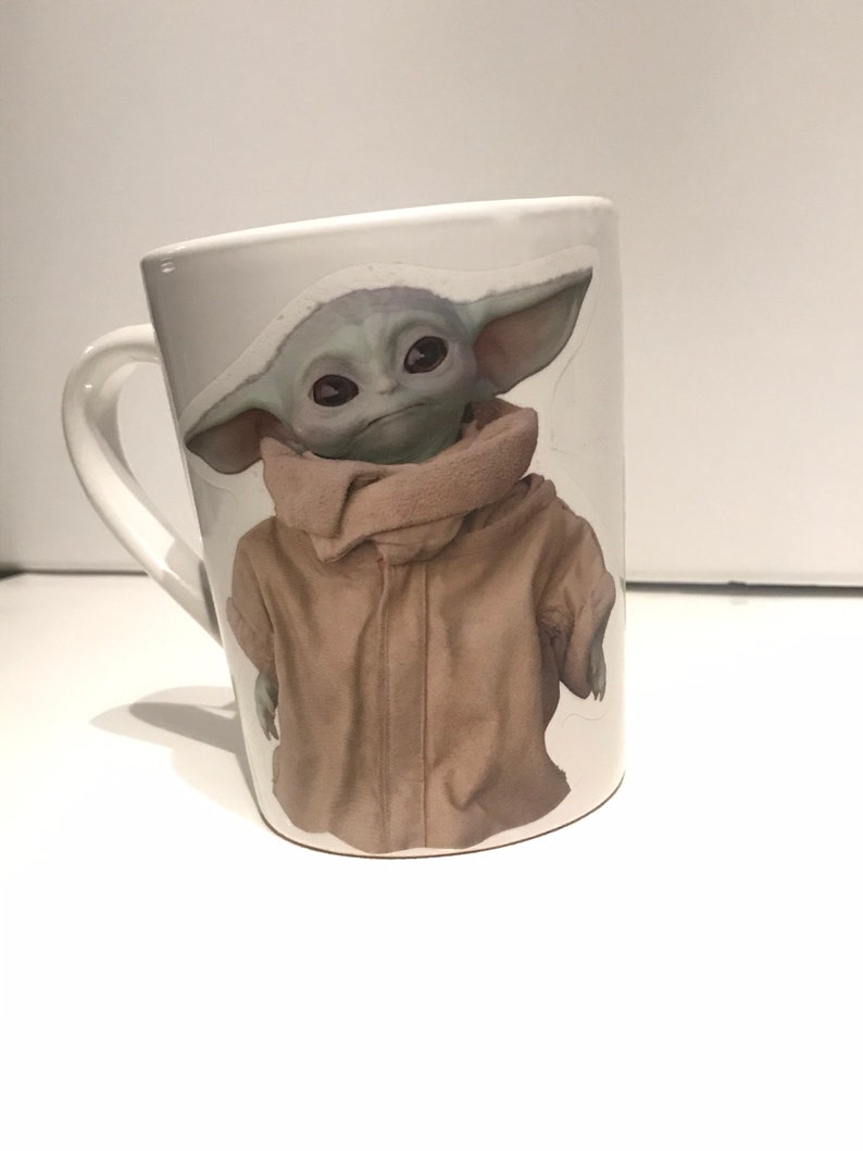 Puede incluir: Taza de cer&aacute;mica blanca con una imagen colorida de un personaje de la franquicia Star Wars, The Mandalorian, conocido como "Baby Yoda".
