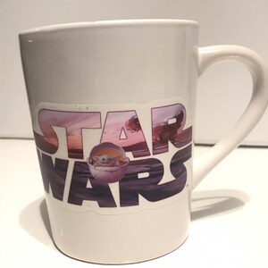 Puede incluir: Taza de cer&aacute;mica blanca con una pegatina de Star Wars que presenta una escena de puesta de sol p&uacute;rpura, rosa y azul con las palabras "Star Wars" en negro y una peque&ntilde;a imagen de Baby Yoda en una c&aacute;psula flotante.