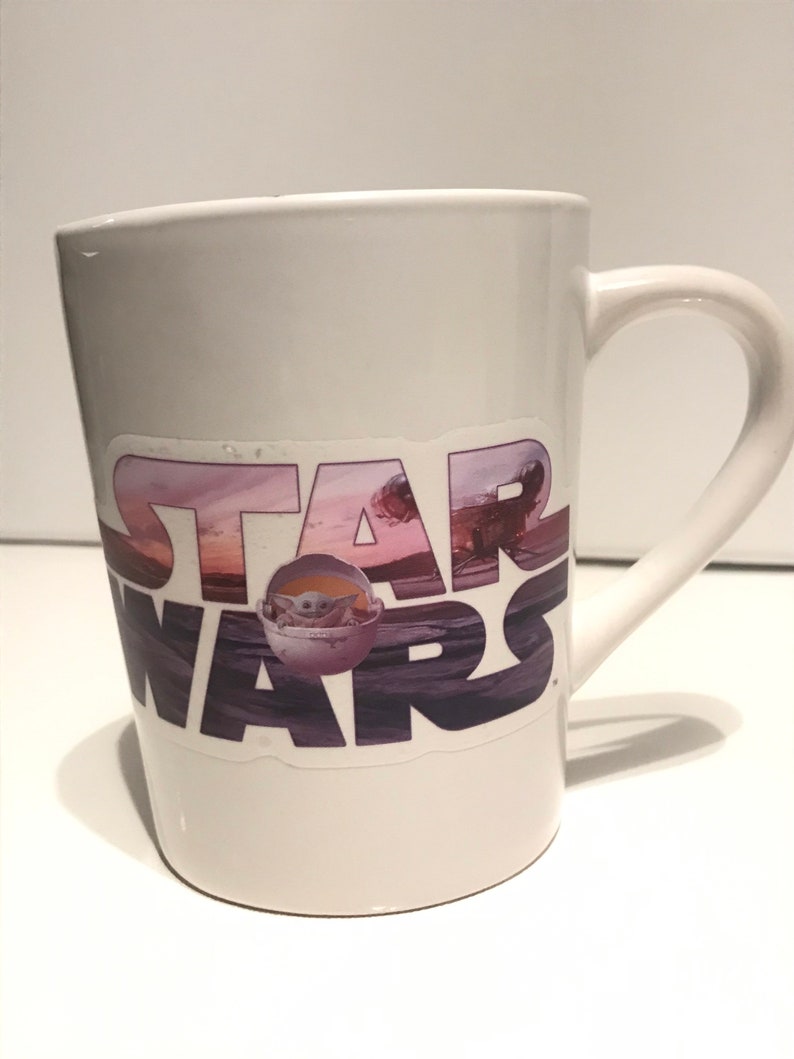 Puede incluir: Taza de cer&aacute;mica blanca con una pegatina de Star Wars que presenta una escena de puesta de sol p&uacute;rpura, rosa y azul con las palabras "Star Wars" en negro y una peque&ntilde;a imagen de Baby Yoda en una c&aacute;psula flotante.