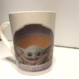 Puede incluir: Taza de cer&aacute;mica blanca con un gr&aacute;fico de un personaje alien&iacute;gena verde de la franquicia de Star Wars, "The Mandalorian". El personaje lleva una t&uacute;nica marr&oacute;n y est&aacute; sentado en una c&aacute;psula redonda con un panel de control.
