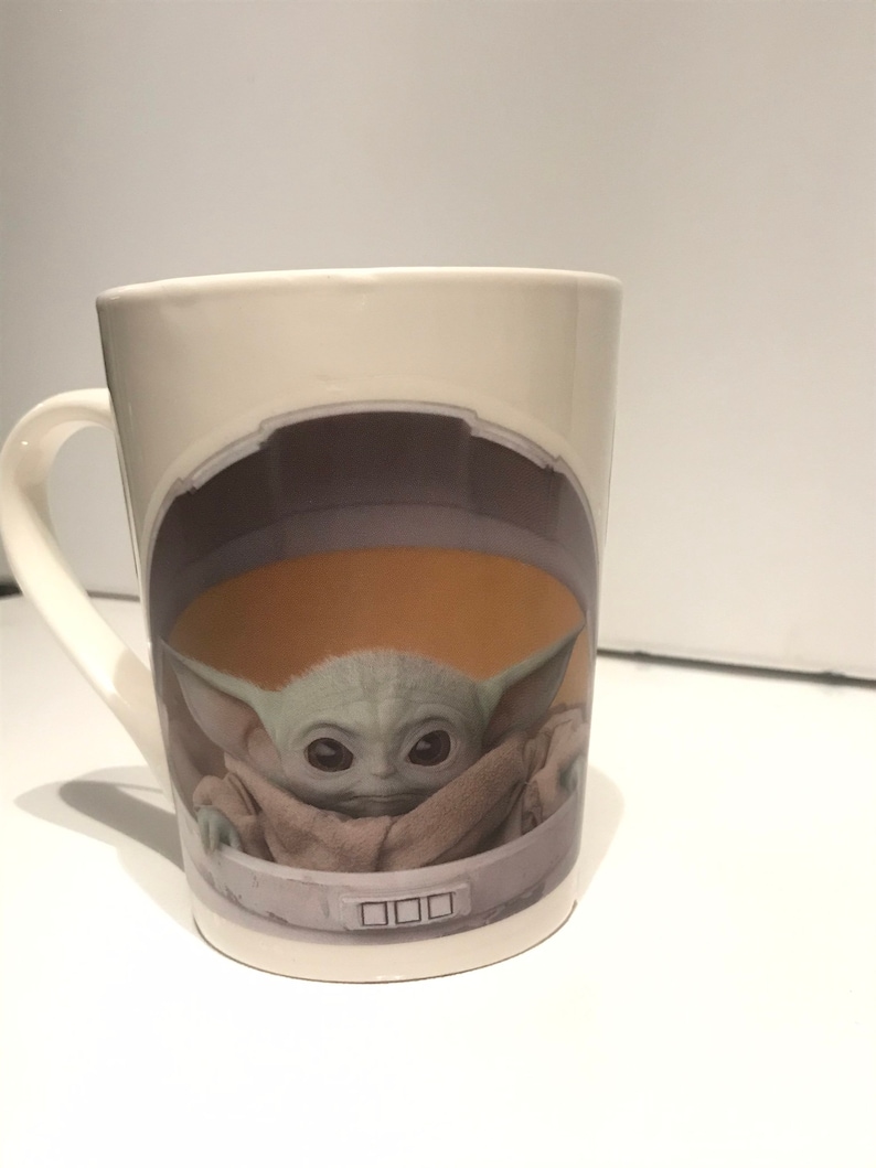 Puede incluir: Taza de cer&aacute;mica blanca con un gr&aacute;fico de un personaje alien&iacute;gena verde de la franquicia de Star Wars, "The Mandalorian". El personaje lleva una t&uacute;nica marr&oacute;n y est&aacute; sentado en una c&aacute;psula redonda con un panel de control.