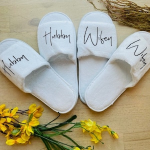 May include: White slippers with the words "Hubby" and "Wifey" written on them. The slippers are on a wooden surface.