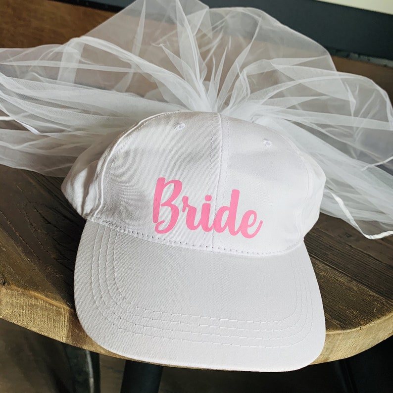 May include: White baseball cap with a pink "Bride" text and a white tulle veil attached to the top.