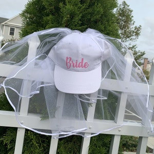 May include: White baseball cap with a white tulle veil attached. The cap has the word "Bride" written in pink cursive letters.