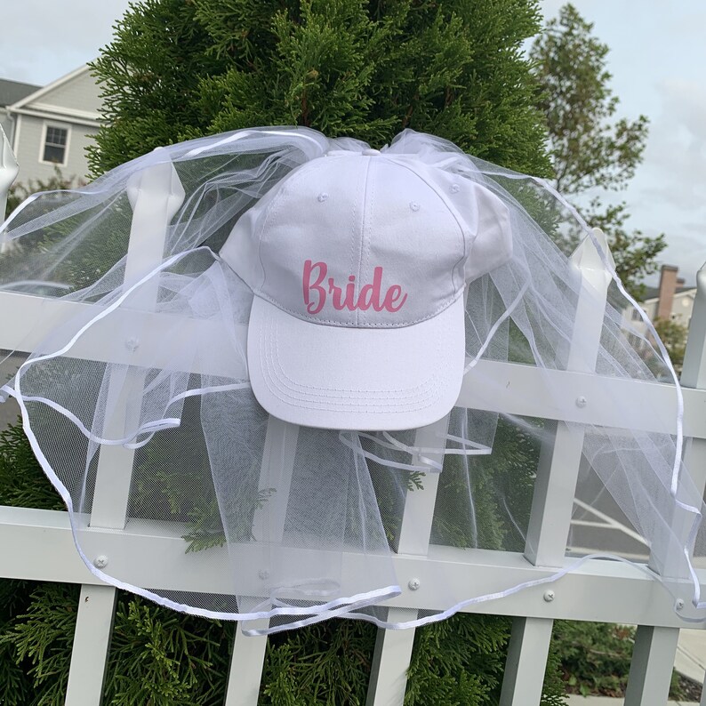 May include: White baseball cap with a white tulle veil attached. The cap has the word "Bride" written in pink cursive letters.